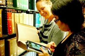 Two women looking at a book and ipad in a library book aisle