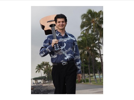 Color of image of slack key guitarist Jeff Peterson with guitar on his back standing in a waterfront park