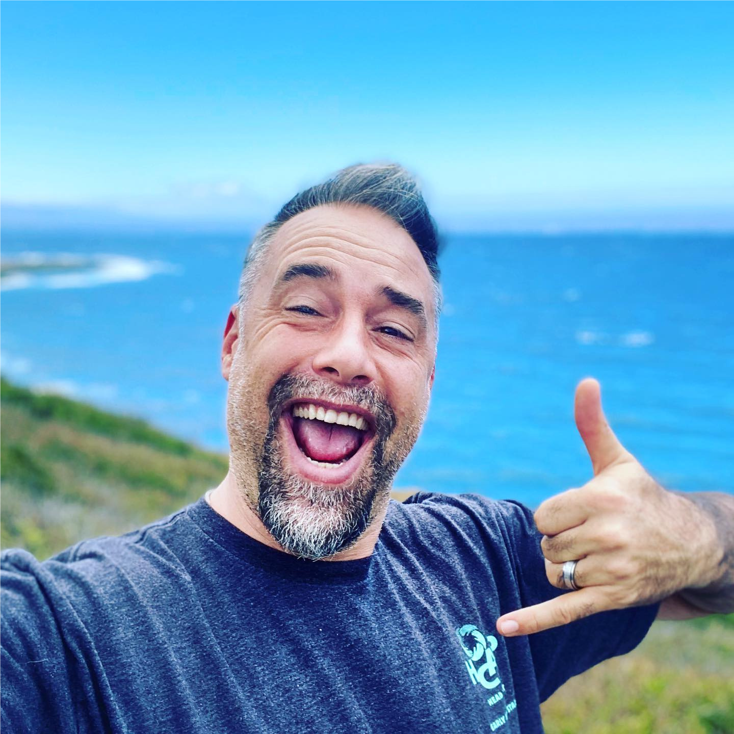 Man in blue shirt with ocean behind smiling and holding up a shaka