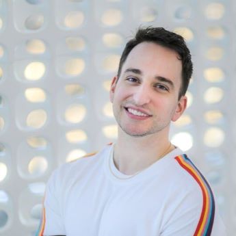 Author Ryan La Sala smiling in a white shirt with a rainbow stripe down their sleeve.