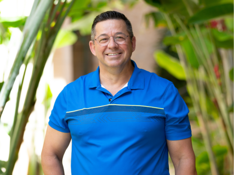 Ron Cahoon in a blue polo smiling at the camera in front of palm leaves.