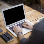 A laptop sitting on a wooden table with a smart phone and a datebook and pen to the left of it. A man is typing on the laptop.