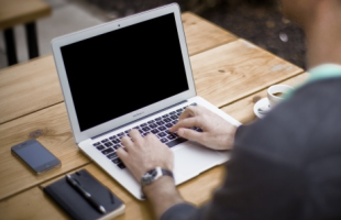 A laptop sitting on a wooden table with a smart phone and a datebook and pen to the left of it. A man is typing on the laptop.