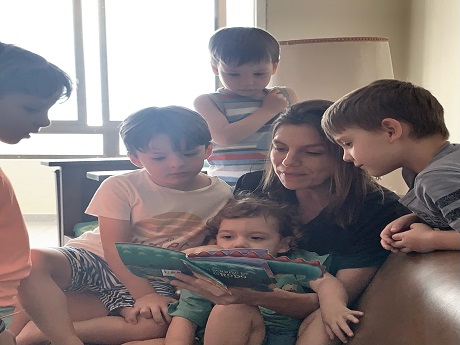 Woman reading a book to several children close to her with a baby on her lap.
