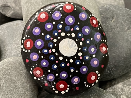 A black river stone sitting on top of gray river stones. The black stone has a mandala pattern of blues, reds, purples, pinks, and white painted on it.