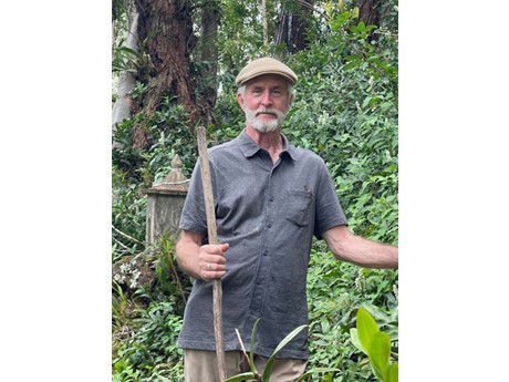 Man with white beard and grey cap holding a walking stick in front of a green hillside.